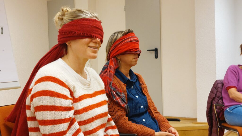 Zwei Teilnehmerinnen des Workshops sitzen auf orangefarbenen Stühlen. Sie haben beide die Augen mit roten Schals verbunden und wirken konzentriert. Beide sind blond, eine trägt einen quergestreiften, weiß-roten Pullover, die andere ein dunkelblaues Hemd mit orangefarbener Strickjacke darüber.